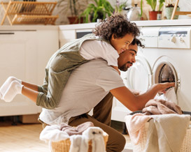 Side view of black child in casual clothes with curly hair smiling and embracing dad loading washing machine during household routine in morning at home.