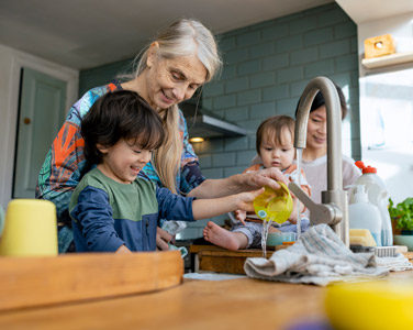 A side-view shot of a mature grandmother standing in the kitchen with her two young grandsons, they are wearing casual clothing and washing cutlery together. The boys mother is besides them drying up.